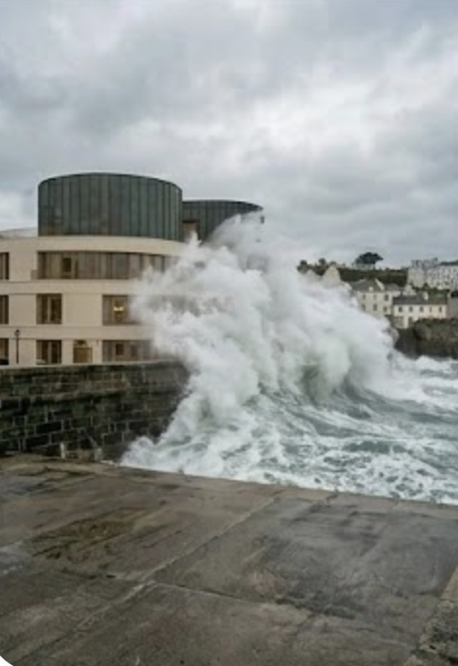 A reference image showing a curved-massing contemporary building with a bronze atrium tower, sitting on a granite Victorian sea wall, a large wave breaking against the wall in front of it on a grey day, with the residential terraces of St Peter Port rising up the cliff behind